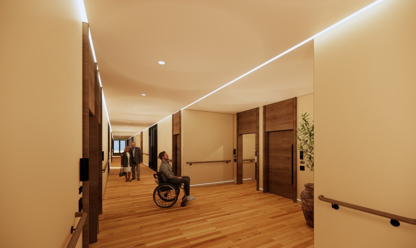A man in a wheelchair in a wide residential corridor with handrails on both sides, a couple walking beyond him